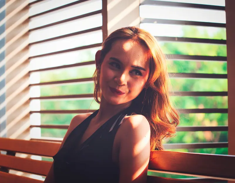 A woman sitting indoors with sunlight filtering through blinds, creating a warm and relaxed ambiance.