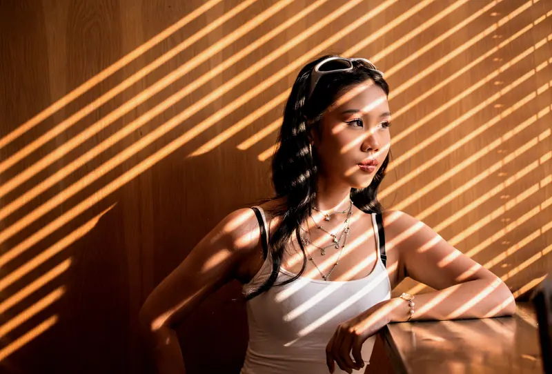 A woman poses elegantly under striped sunlight from window blinds indoors.