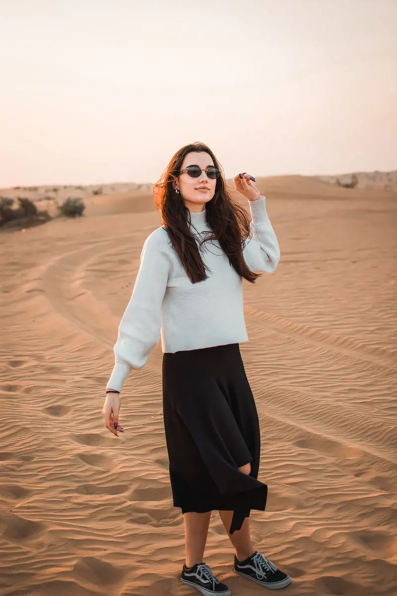 A young woman strolls through sunlit desert dunes, wearing sunglasses and casual attire.
