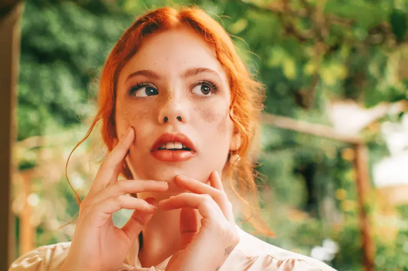 Close-up portrait of a thoughtful redheaded woman in a serene outdoor setting with greenery.