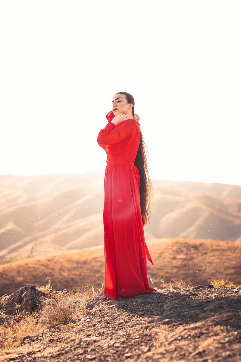 A woman in a flowing red dress stands gracefully in a sunlit desert landscape.
