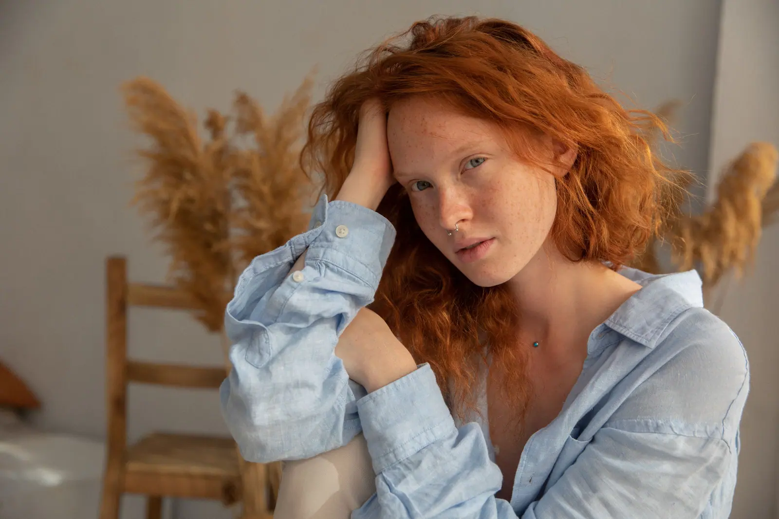 Woman with long copper waves in a soft blue shirt, sitting beside pampas grass in a sunlit adobe-toned room