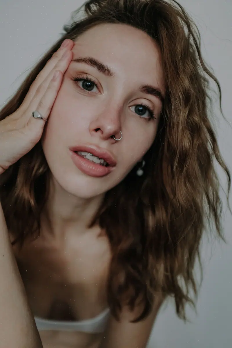 Close-up portrait of a young woman with wavy hair and nose piercing against a white background.