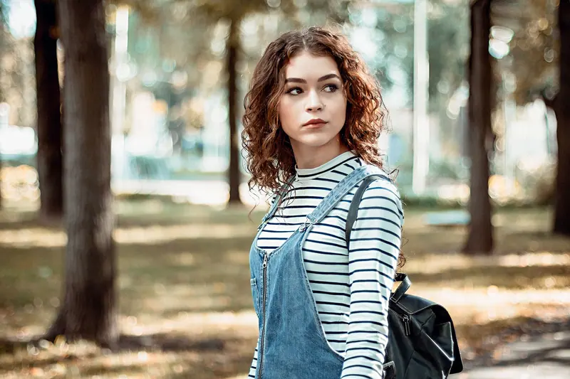 A fashionable woman with curly hair in a striped shirt poses outdoors in a Vilnius park.