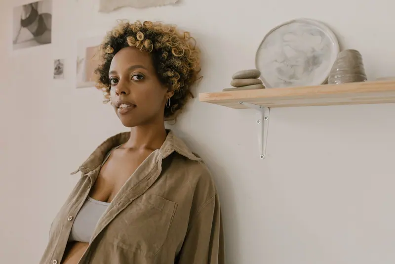 Stylish woman with curly dyed hair leaning against a wall in a peaceful indoor setting.