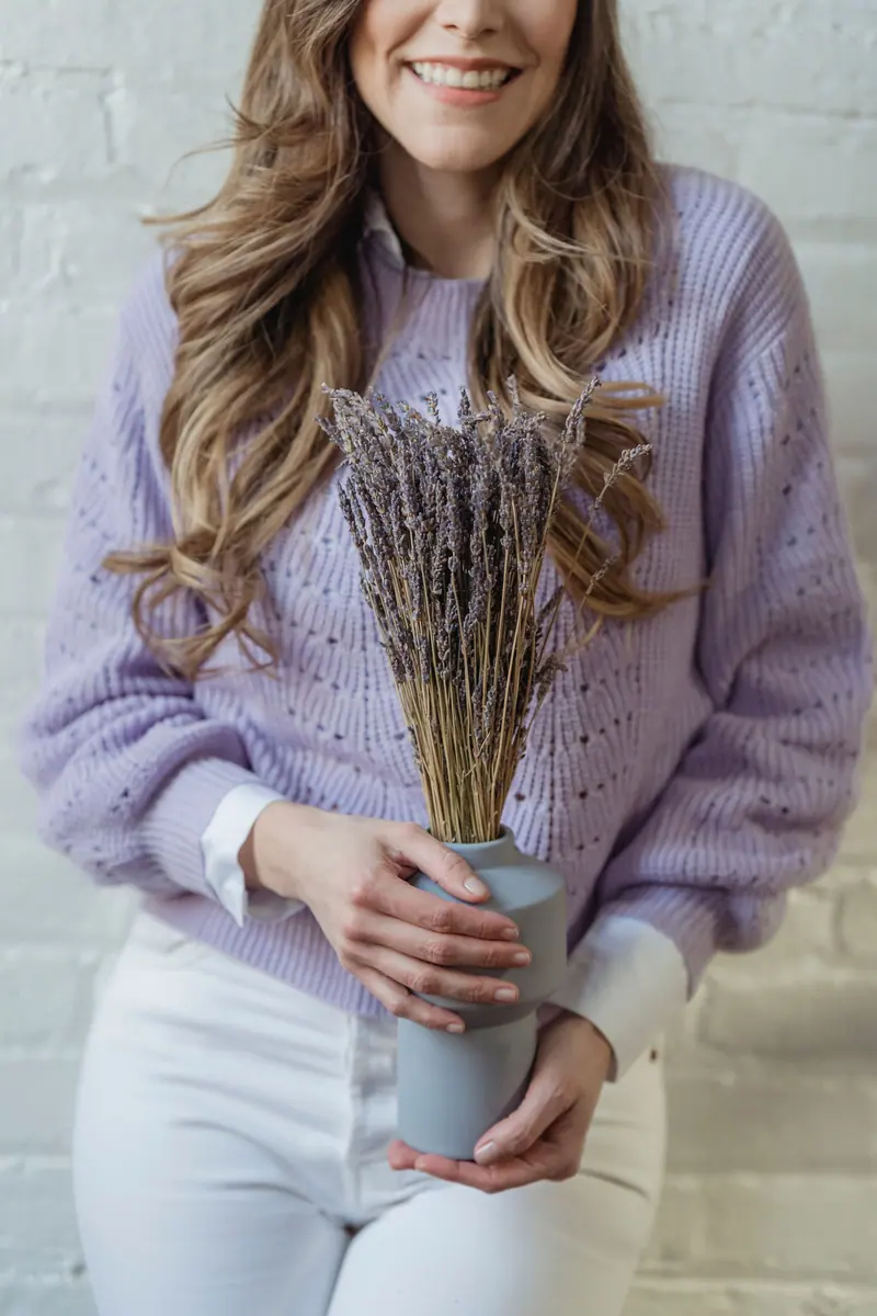 Smiling woman holding lavender bouquet in vase, wearing a purple sweater against a brick wall.