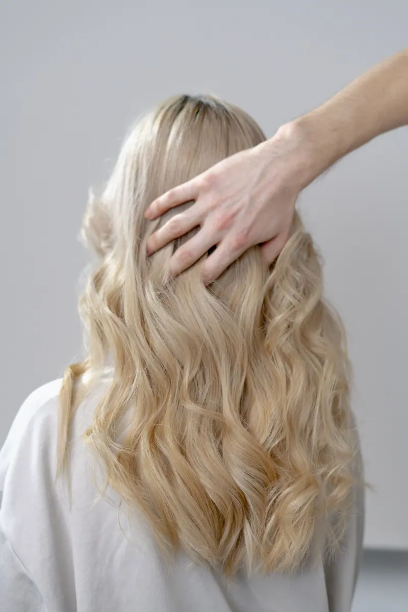 A stylist's hand touches the long, blonde curly hair of a woman from behind.