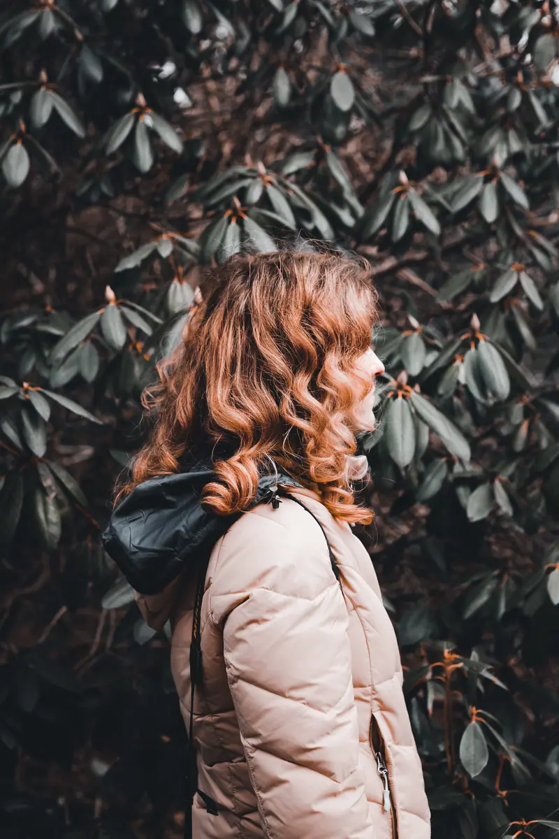 A red-haired woman in a warm jacket stands amidst green shrubs, embracing nature's tranquility.