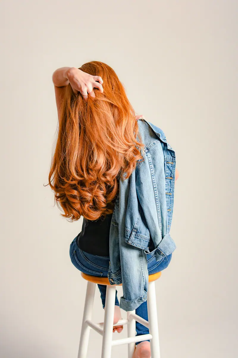 A woman with red hair sits on a stool wearing denim jacket, showcasing her hair.