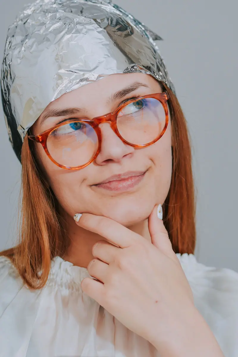Woman with red glasses and tinfoil hat in a thoughtful pose indoors.