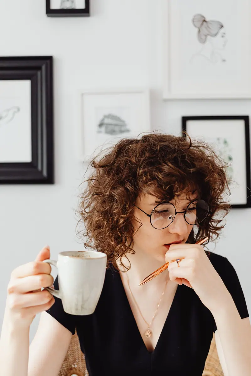 Curly-haired woman with eyeglasses holding a coffee cup and pen, thinking in a modern indoor setting.