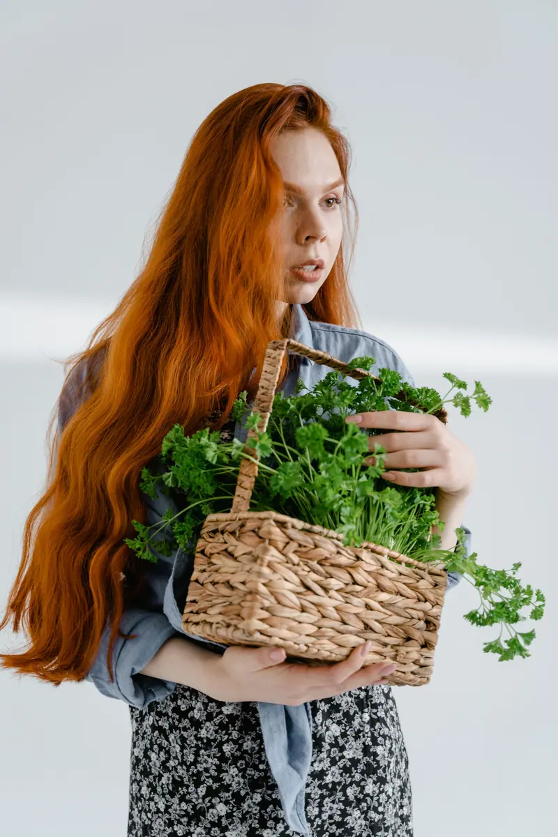 Portrait of a woman with auburn hair holding a wicker basket full of green parsley indoors.