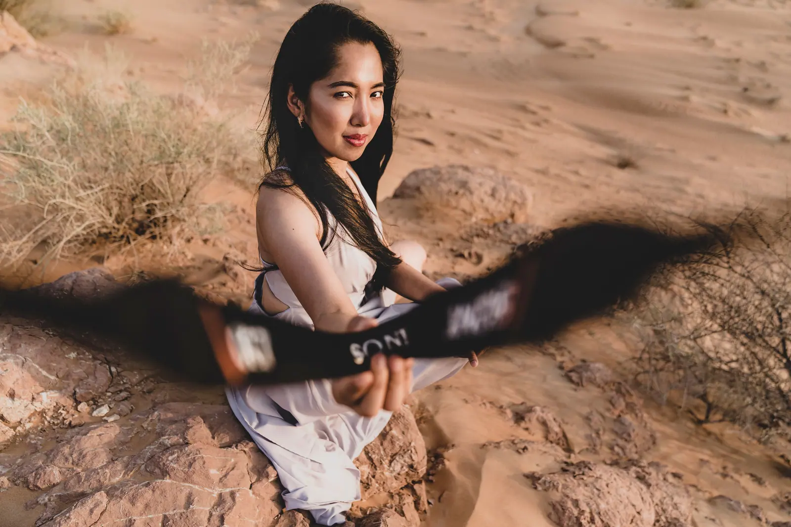 Woman sitting on rocks in a desert, holding a camera strap, with focused gaze and sandy background.