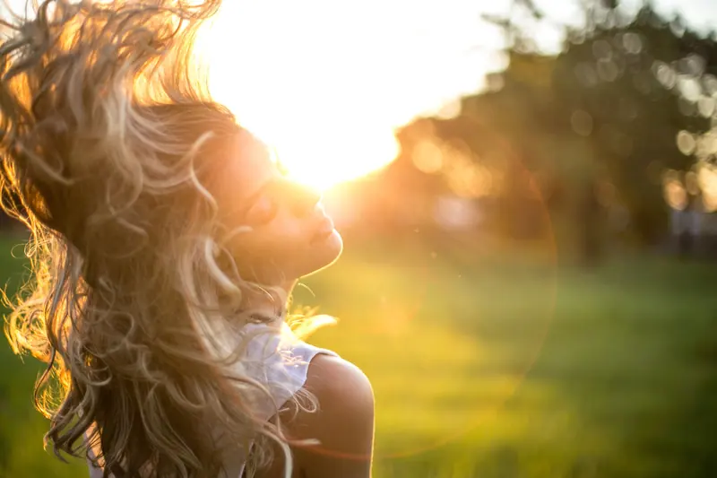 A young woman tosses her long, wavy blond hair in the sunlight during golden hour outdoors.