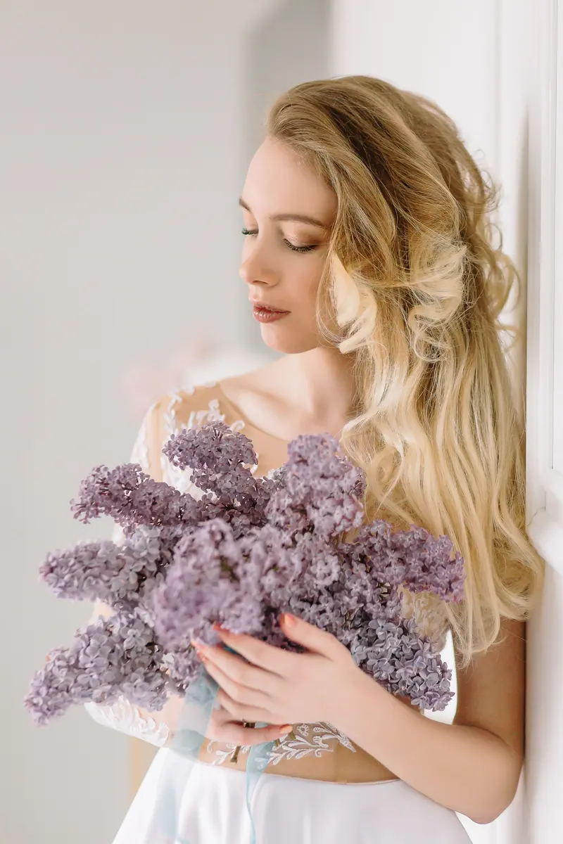 A beautiful bride with blonde hair holding a bouquet of purple flowers in a serene indoor setting.
