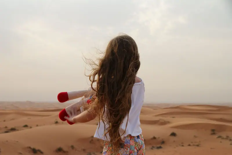 A young girl with long hair holding a toy stands in a vast desert landscape.