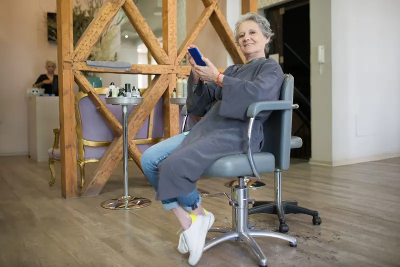 Elderly woman seated in a hair salon chair using a cellphone, relaxed indoor setting.