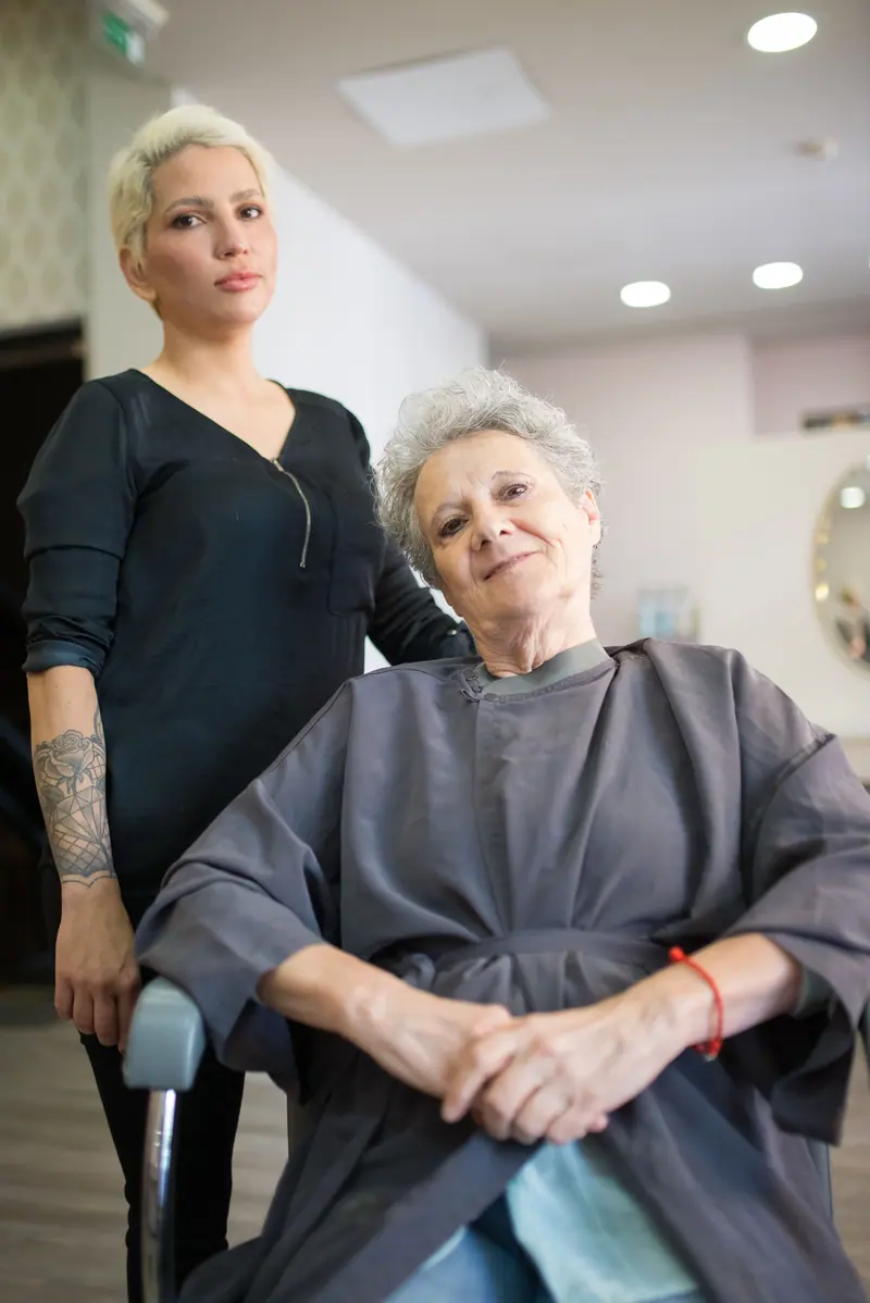 An elderly woman receives a haircut at a salon in Portugal, with a stylist standing beside her.
