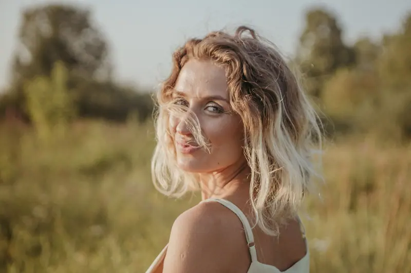 A smiling woman with blond hair glances over her shoulder in a sunlit field.