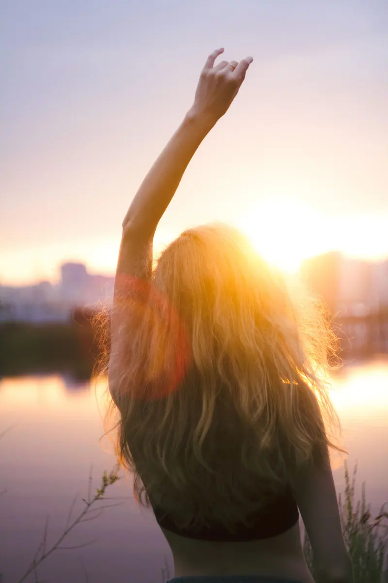 Silhouette of a woman raising her hand at sunset by the water, creating a serene and hopeful atmosphere.