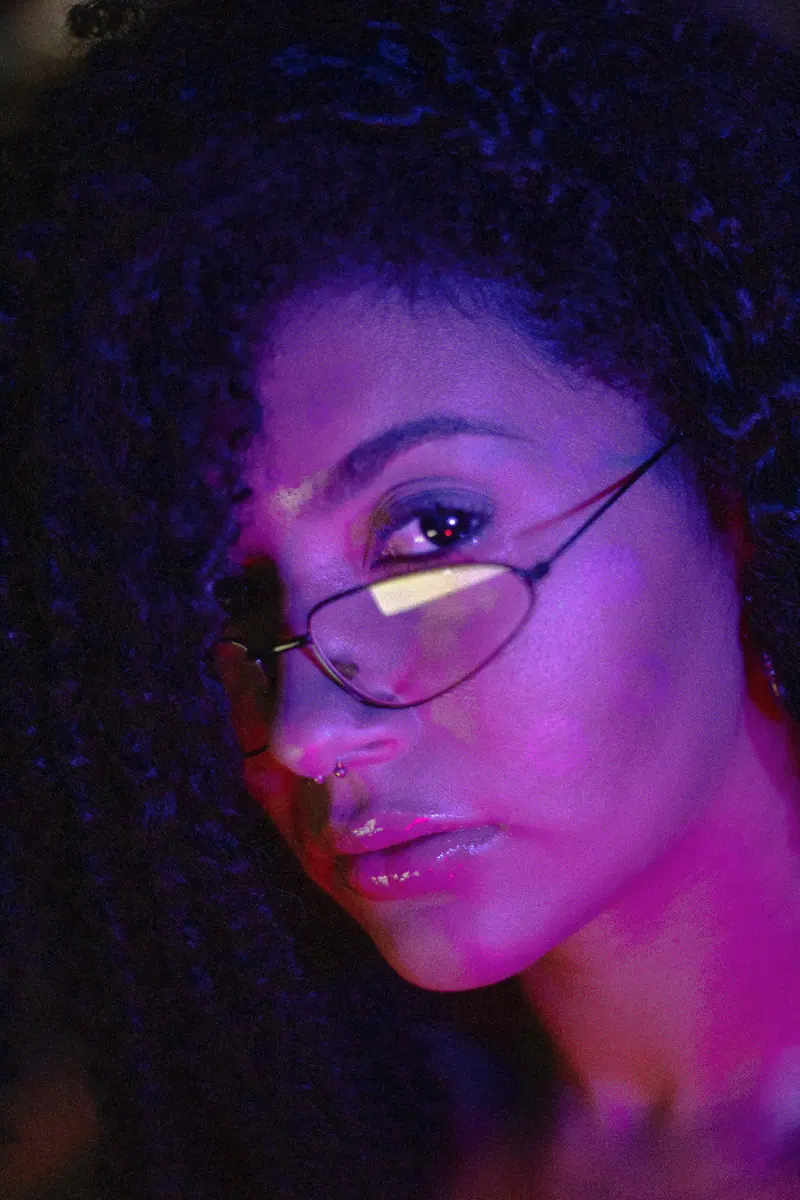 Artistic portrait of a woman with curly hair and glasses, emphasized by vibrant purple lighting.