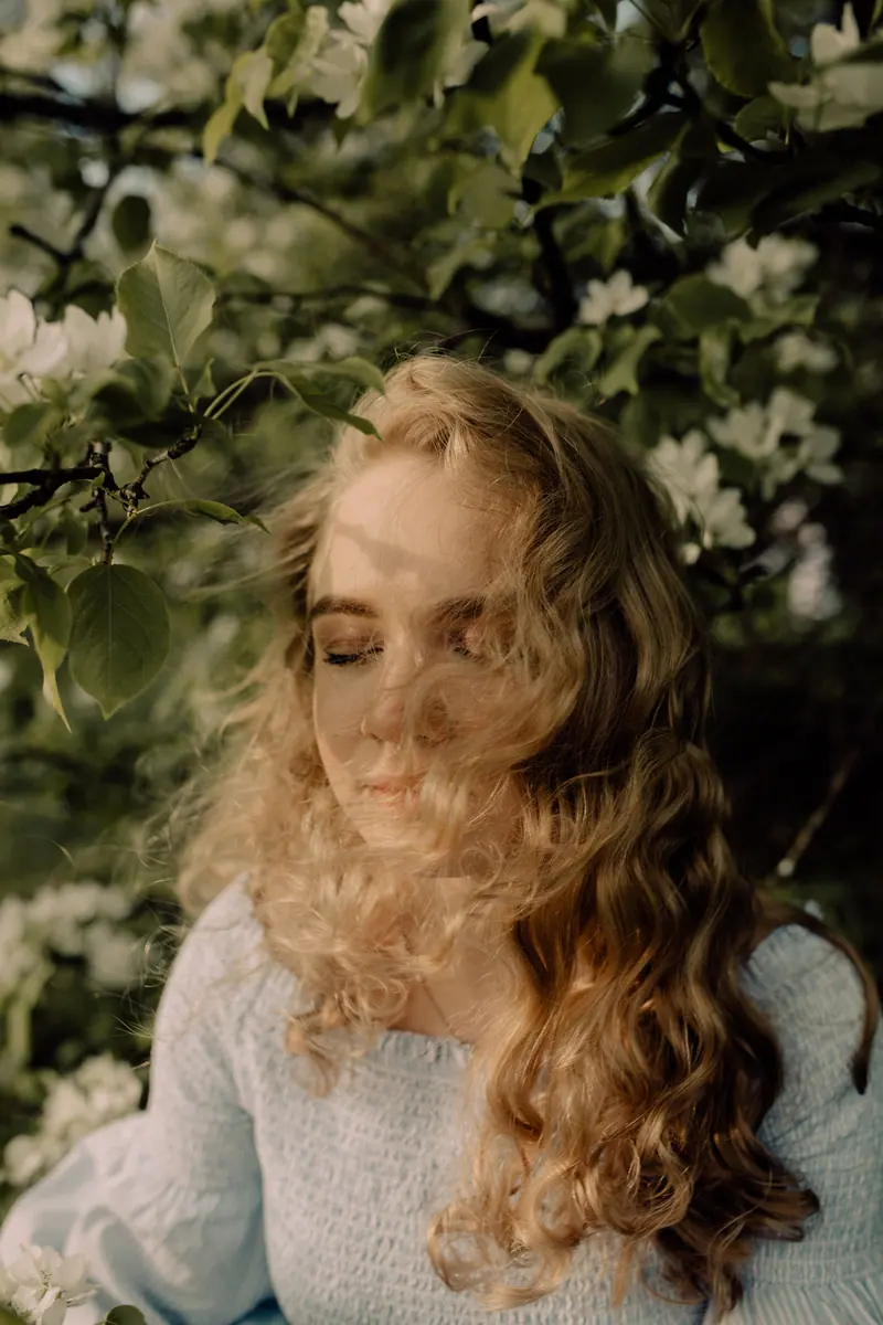 A serene moment of a woman with wavy blonde hair amidst lush green foliage in spring.