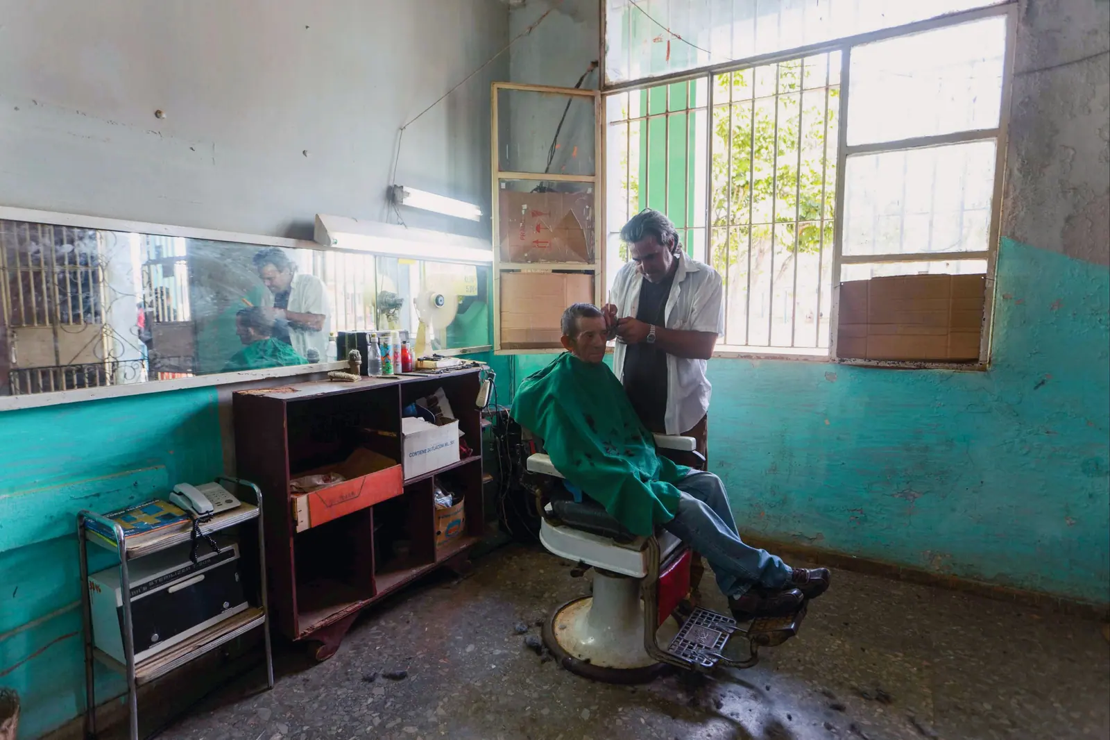 Barber cutting an older man's hair in a classic shop with a green-tiled wall and morning light through tall windows