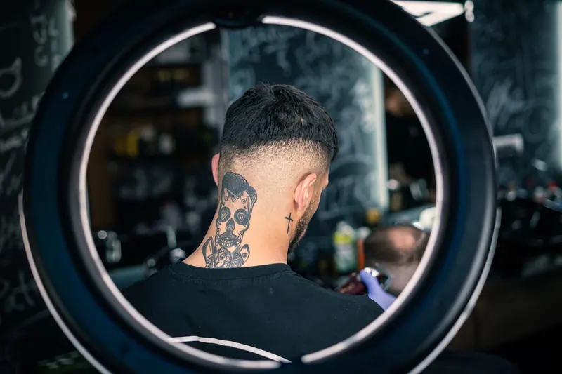 Back view of man with skull neck tattoo getting a haircut surrounded by a ring light in a barbershop.