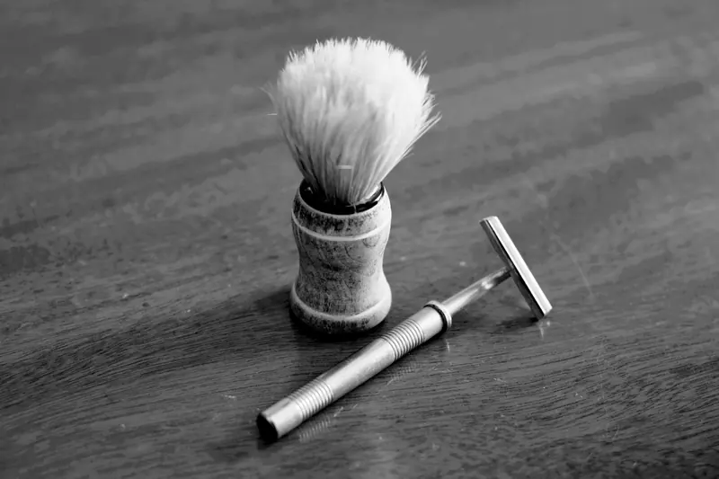 Black and white image of a classic shaving brush and razor on a wood table.