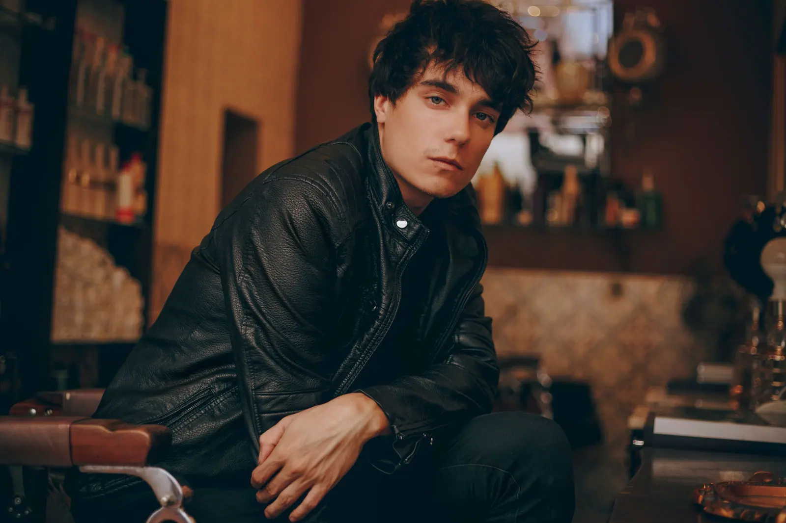 Young man with tousled dark hair in a leather jacket seated in a warm, wood-lined barbershop