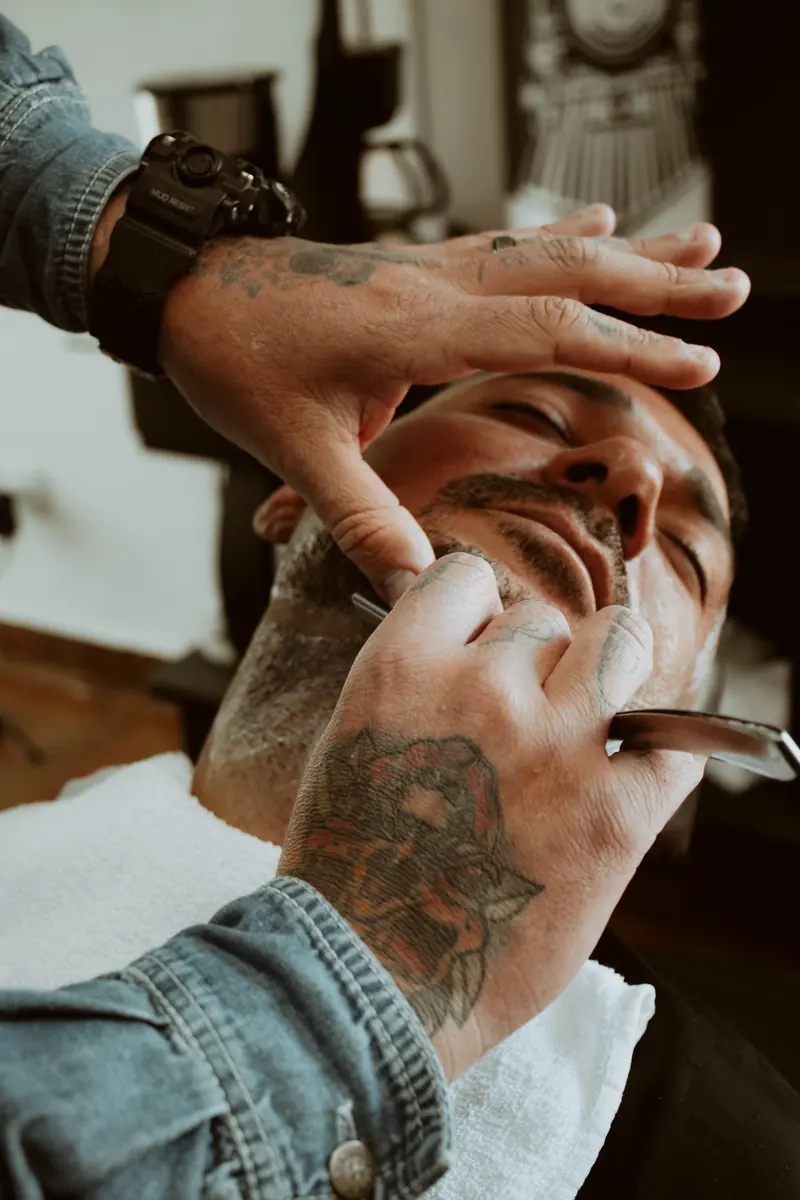 Close-up of a tattooed barber skillfully shaving a client's face in a modern barbershop.