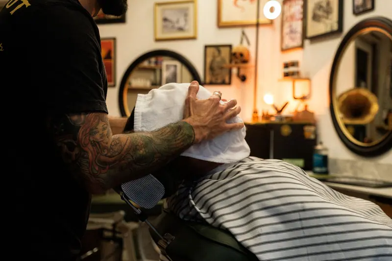 A barber with tattoo sleeves applies a hot towel in a vintage-style barbershop setting.