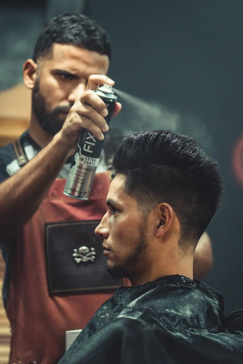 Experienced stylist applies finishing spray to client's hair during grooming session at modern barbershop.