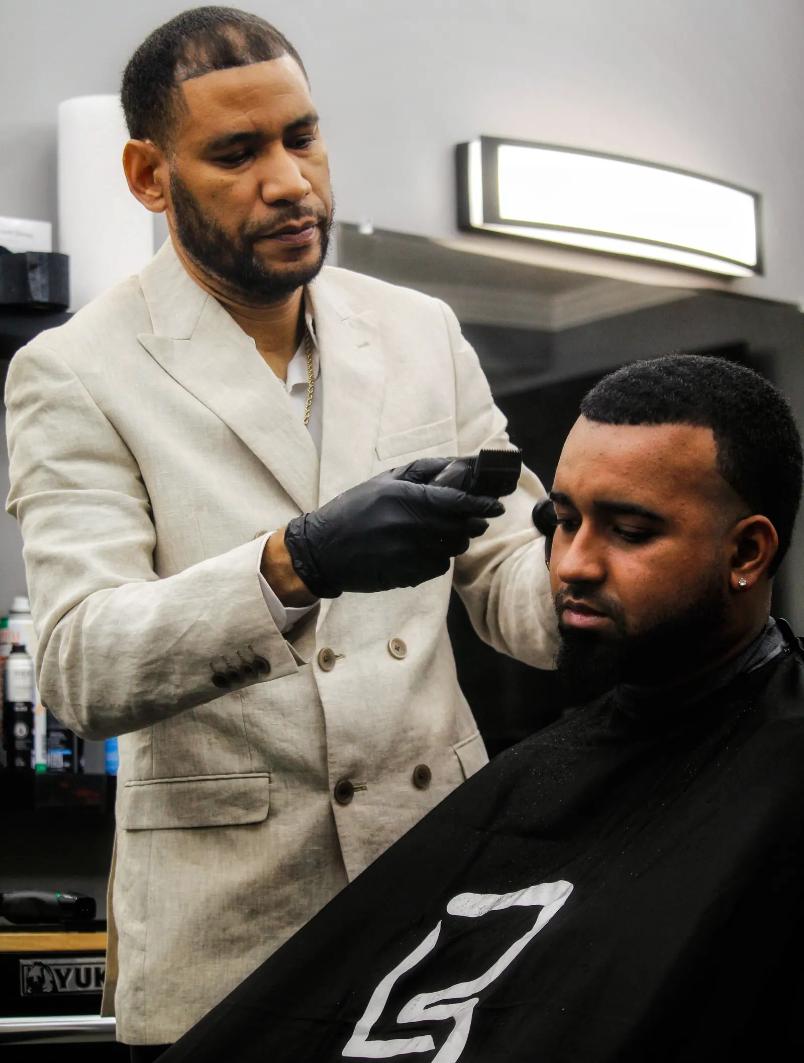 Barber in a cream suit and black gloves lining up a client's beard in a bright shop