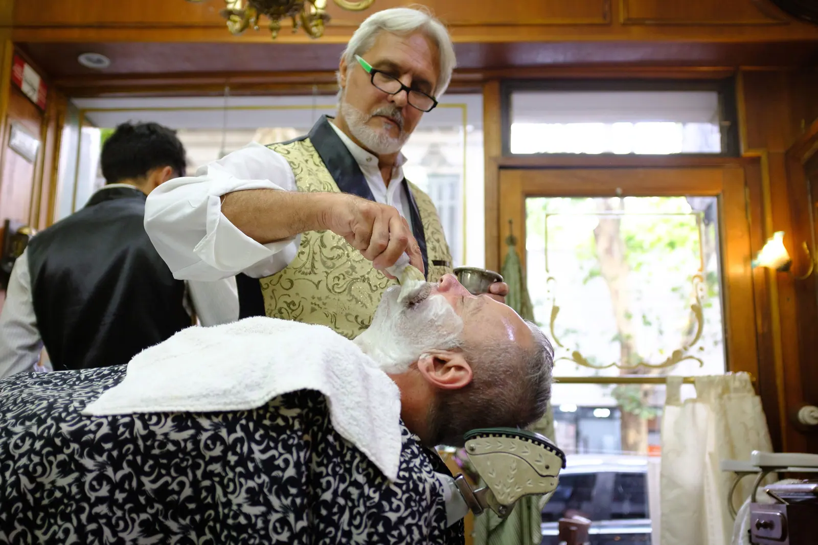 Silver-haired barber giving a client a hot-towel straight-razor shave in a wood-paneled vintage barbershop