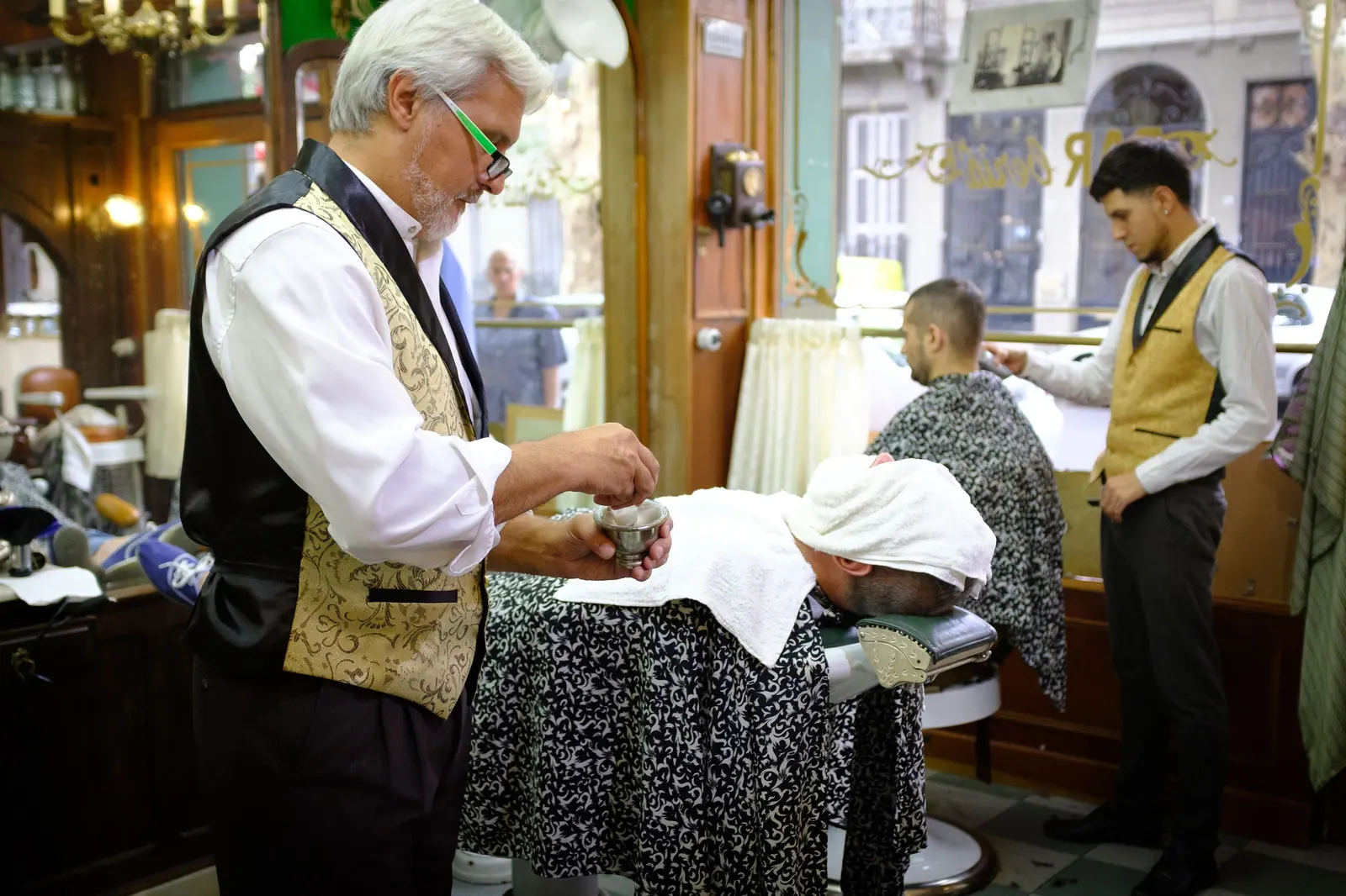 Silver-haired barber in a brocade vest preparing a hot-towel shave in an old-world wood-panelled parlor