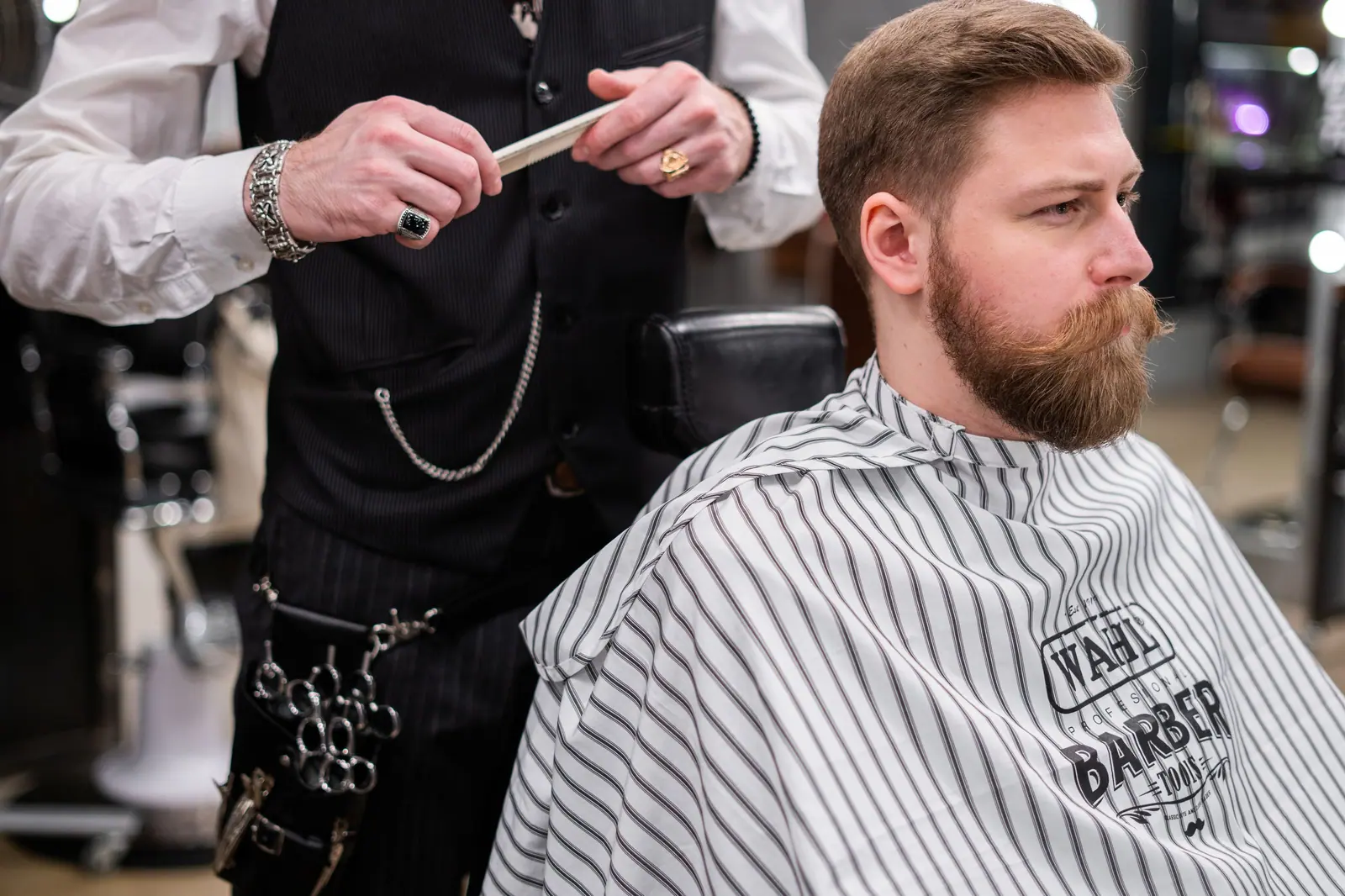 Bearded barber in a waistcoat combing a client's hair in a bright, mirror-lined barbershop