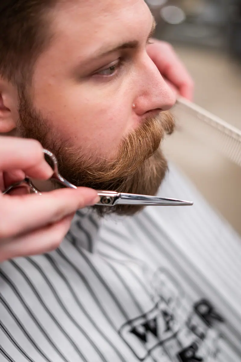 Close-up shot of a barber grooming a bearded man's beard and mustache with scissors.