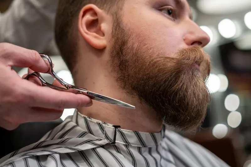 Detailed shot of a barber trimming a man's beard with scissors in a barbershop.