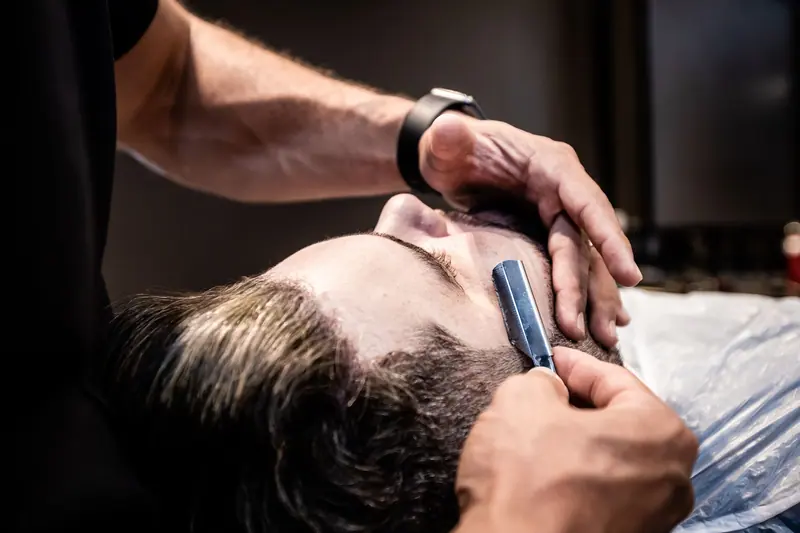 A barber skillfully shaves a client's beard in a modern barber shop setting.