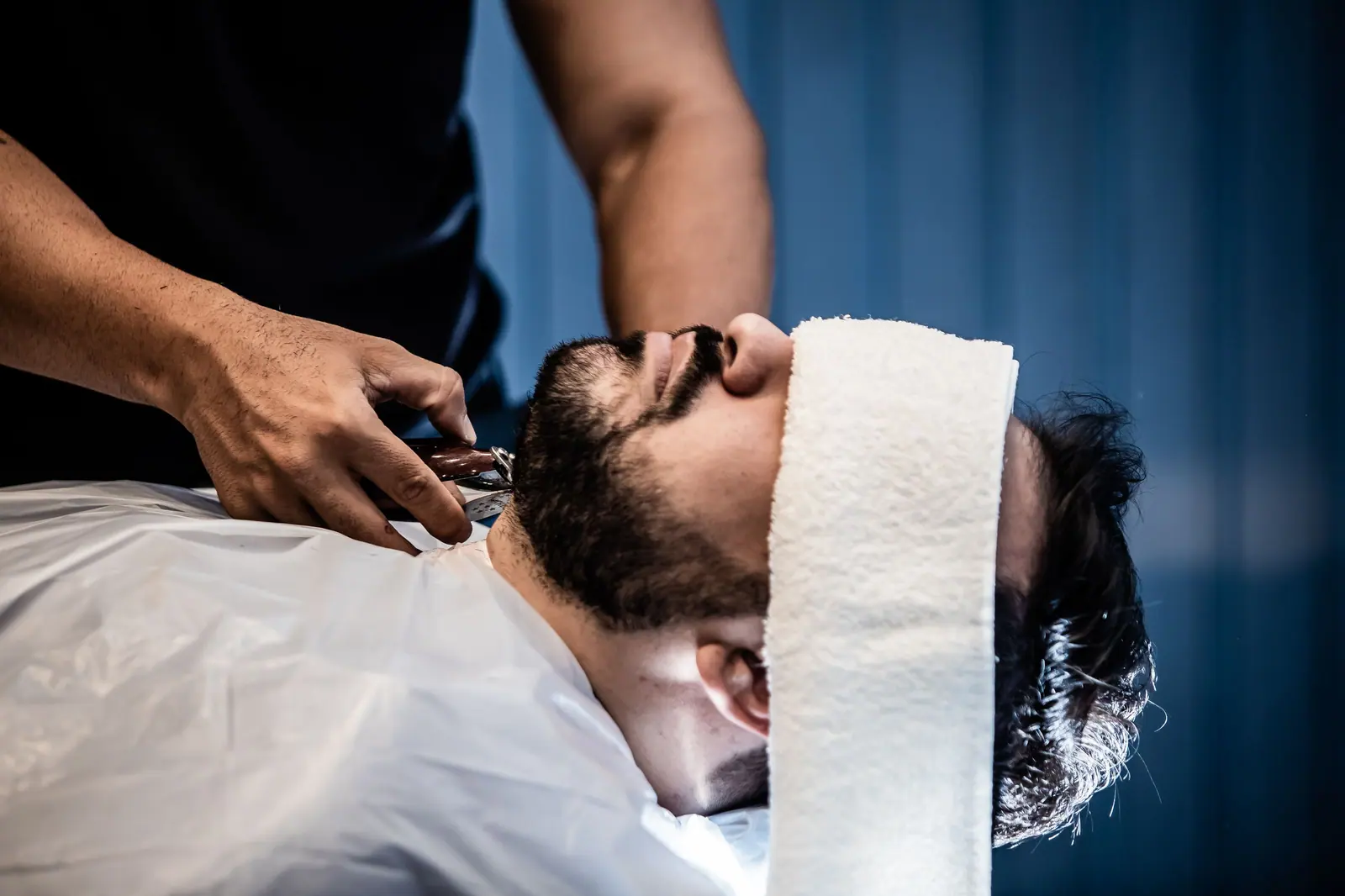 Barber trimming a relaxed client's beard under a hot towel against a navy curtain