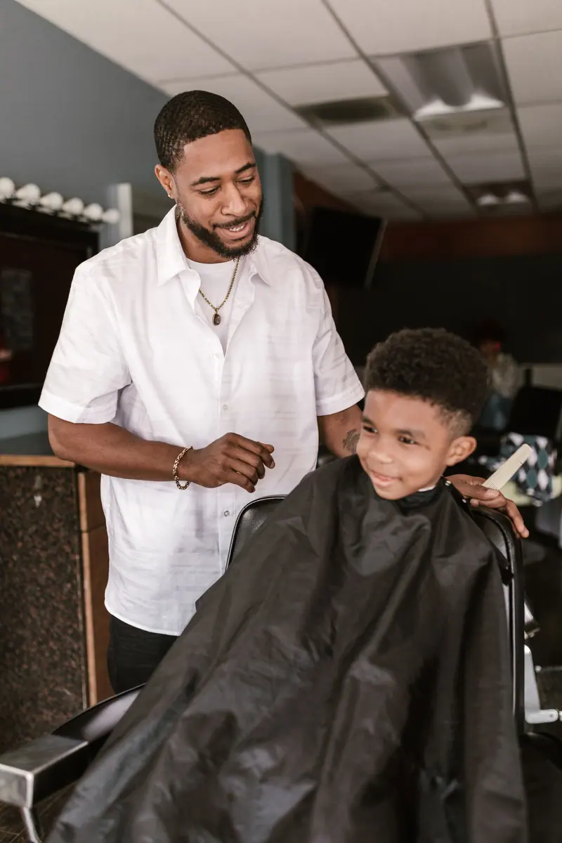 A barber cutting a young boy's hair inside a barbershop. Smiling interactions create a warm atmosphere.