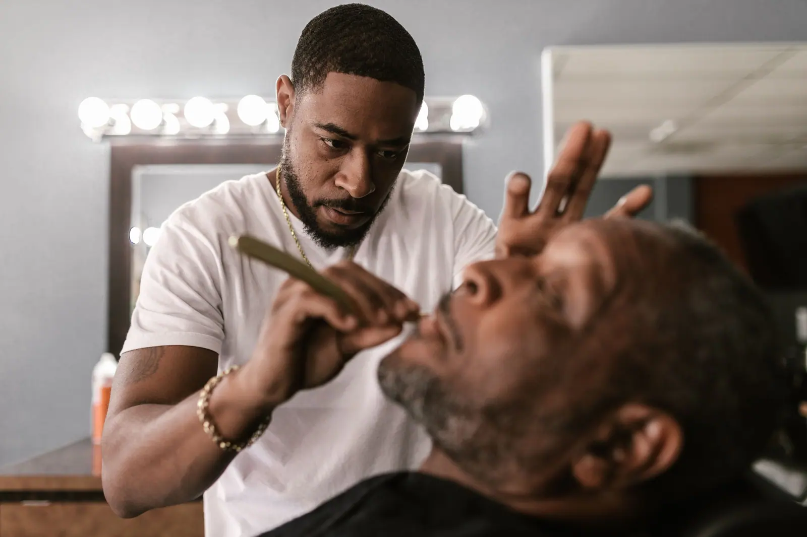 Barber giving a smiling client a straight-razor shave in a bright, mirror-lined barbershop