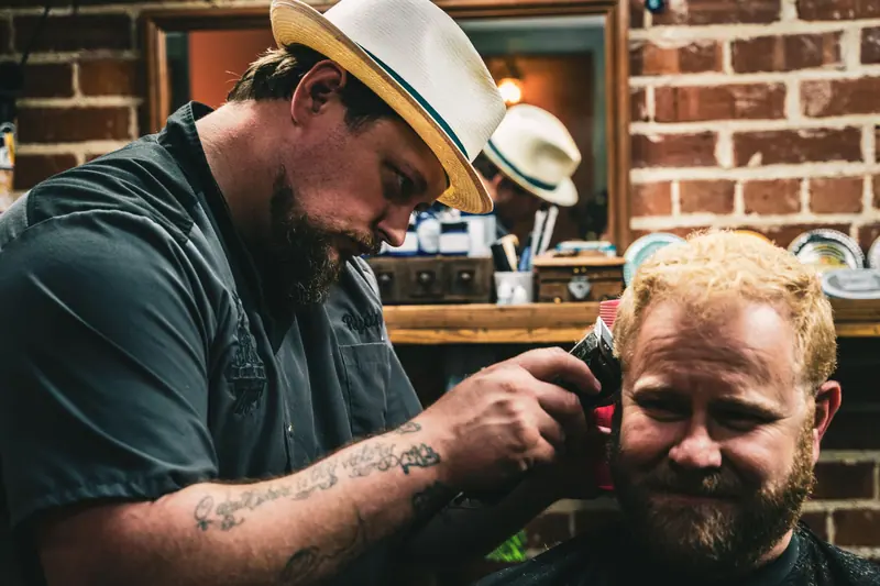 A barber wearing a fedora gives a haircut with a clipper to a bearded man inside a barbershop.
