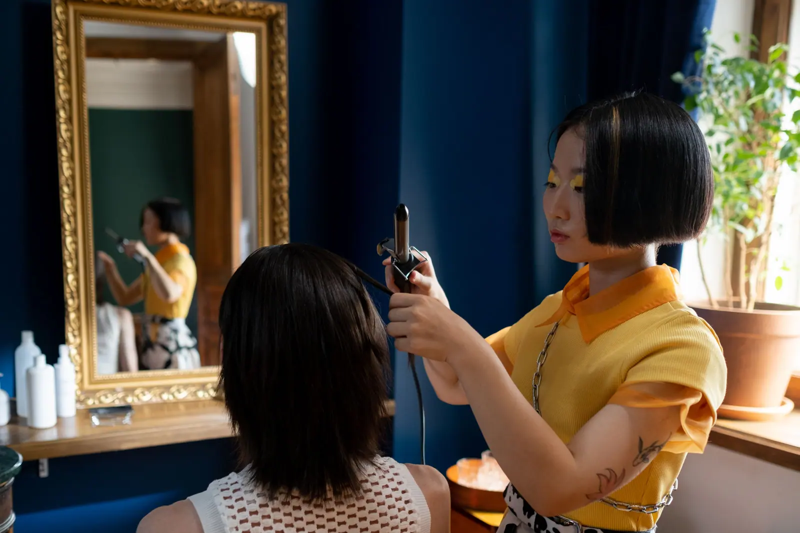 Stylist in marigold finishing a client's sleek bob in a cobalt-blue salon with a gilded mirror
