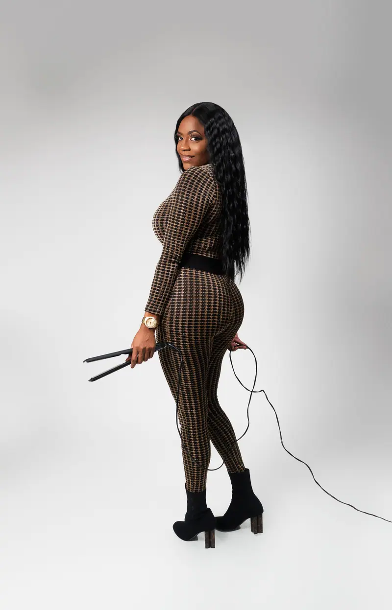 Elegant black woman in chic attire holding a flat iron, showcasing hairstyling skills.