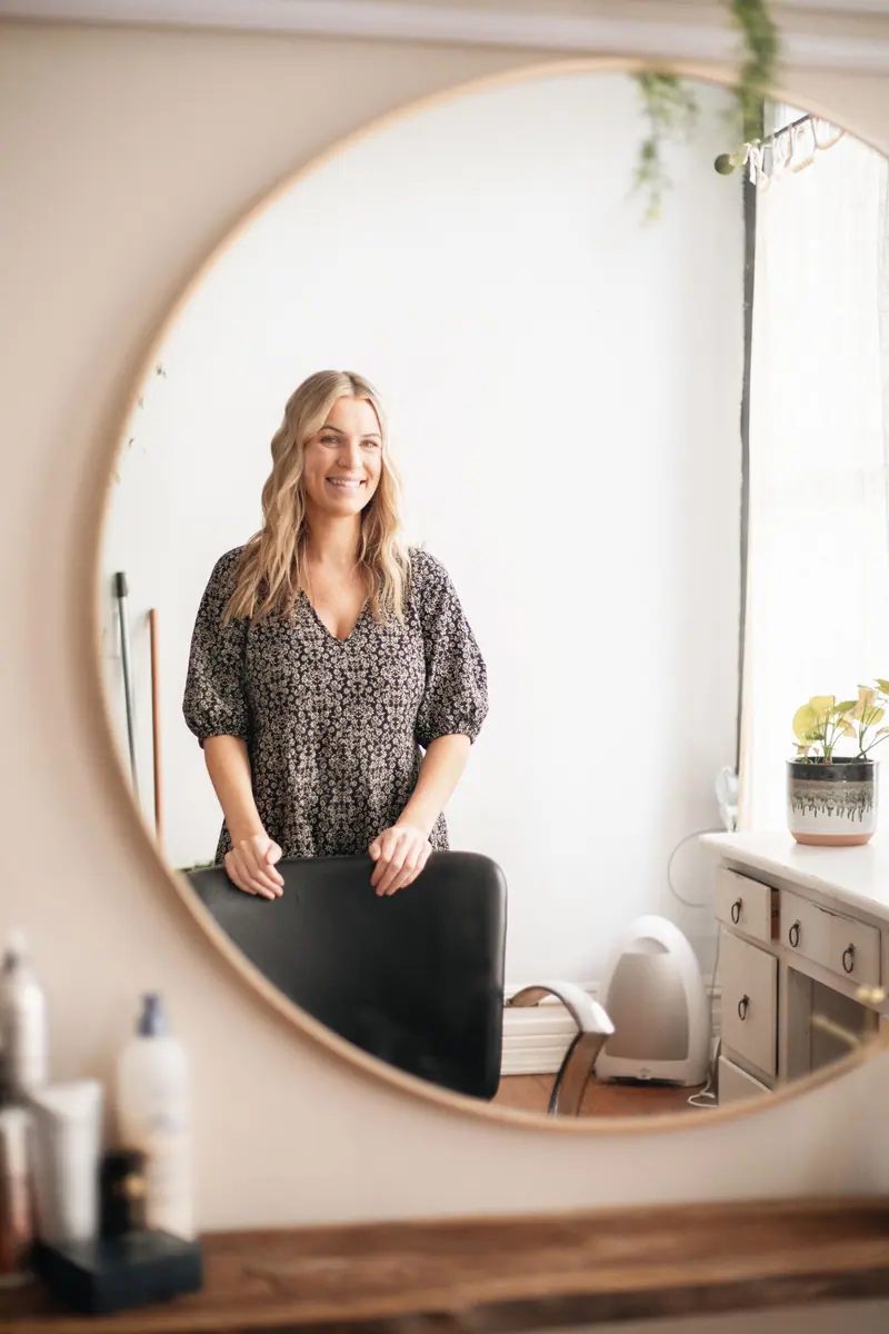 Stylist standing before a round mirror in a bright, plant-filled studio
