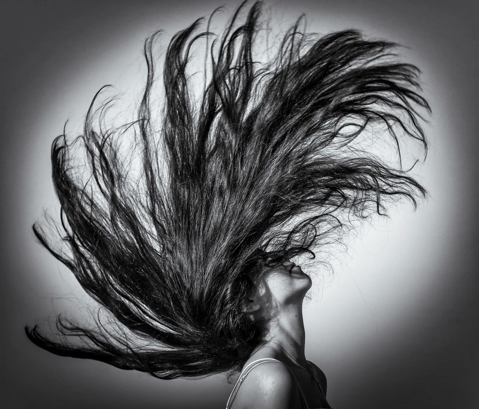 Black-and-white portrait of a woman whipping long hair upward against a grey studio wall
