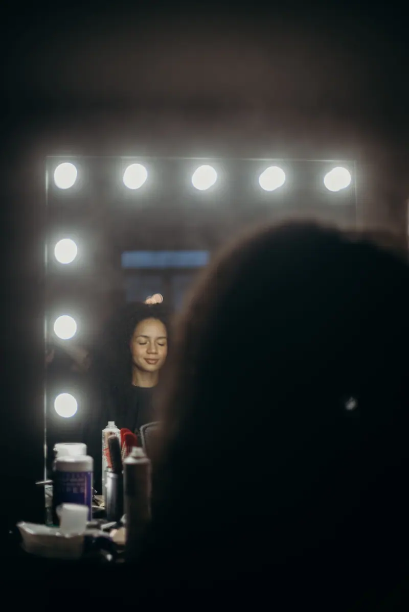 Woman at a bulb-lit vanity mirror in a dark salon