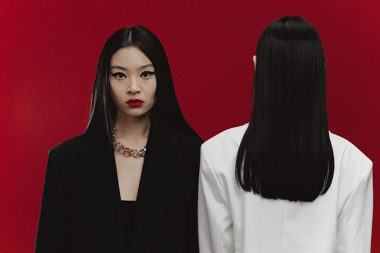 Two women with glass-straight black hair, one seen from behind, against a lipstick-red backdrop
